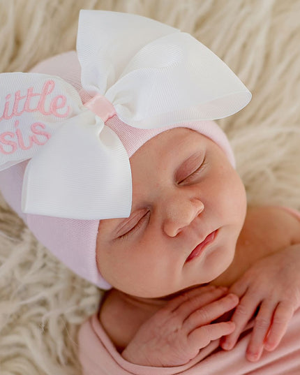 Baby wearing pink hospital hat with oversized white ribbon bow embroidered with 'little sis'