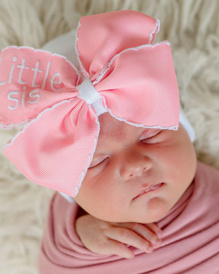 Baby wearing white hospital hat with oversized pink bow that says little sis