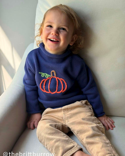 A boy sitting wearing a blue rollneck sweater with a pumpkin design 