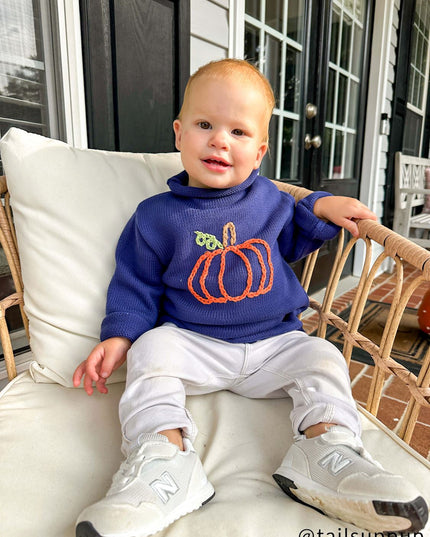 Baby sitting wearing a blue rollneck sweater with a pumpkin design 