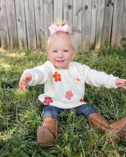 Baby sitting wearing cream sweater with crochet flowers and jeans