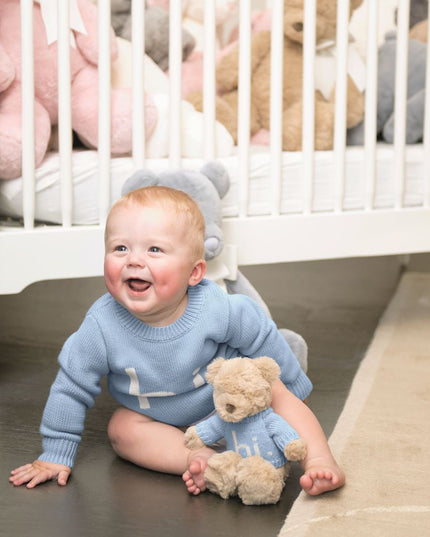 Baby in blue 'hi' sweater sitting on the floor with teddy bear