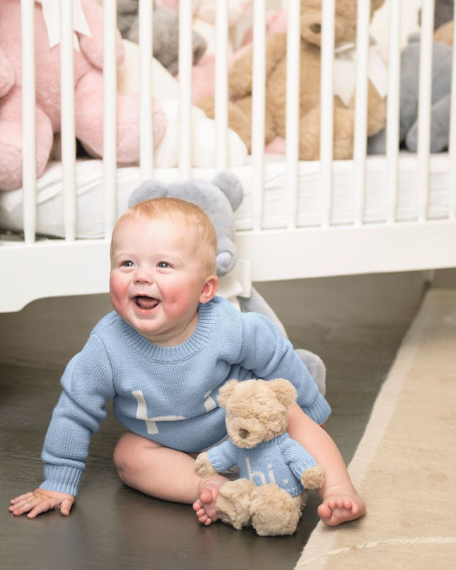 Baby in blue 'hi' sweater sitting on the floor with teddy bear