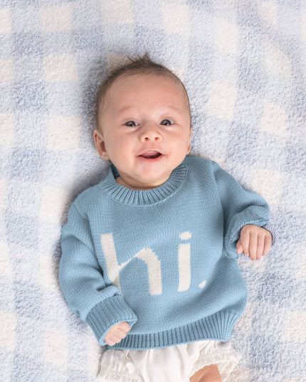 Baby wearing a blue sweater with 'hi' on it, lying on a checkered blanket