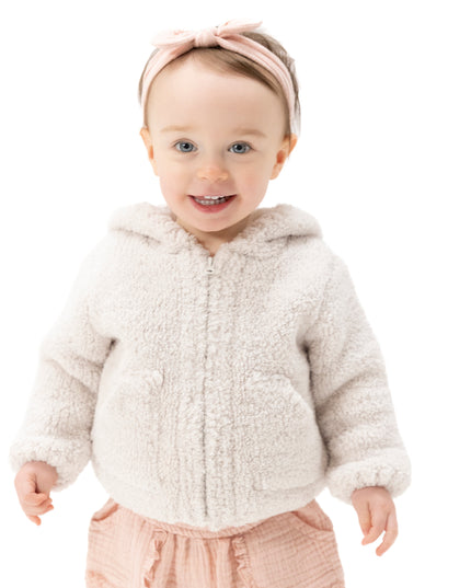 A smiling toddler wearing a cream chenille zip-up hooded jacket, a pink bow headband, and pink pants