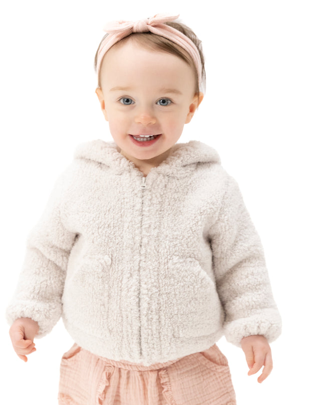 A smiling toddler wearing a cream chenille zip-up hooded jacket, a pink bow headband, and pink pants