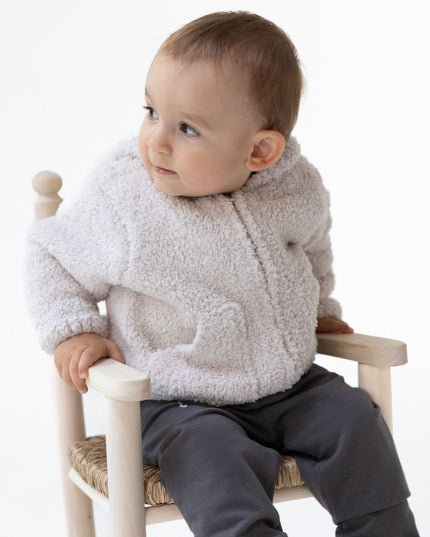 A baby sitting on a small wooden chair wearing an off-white chenille fleece zip-up and dark gray pants