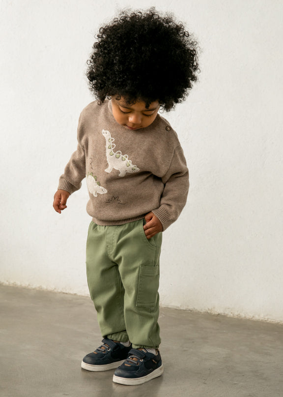 Child wearing a brown sweater with dinosaurs and green pants 