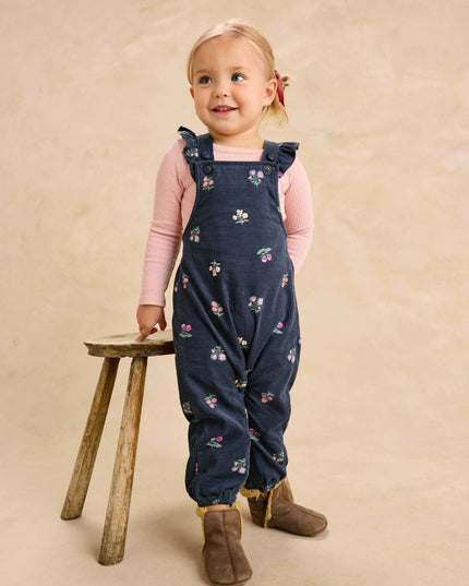 A toddler girl wearing deep midnight corduroy overalls with floral pattern and ruffle straps and a pink undershirt