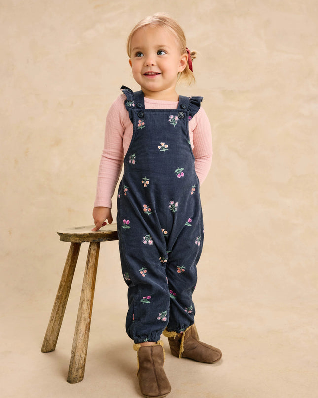 A toddler girl wearing deep midnight corduroy overalls with floral pattern and ruffle straps and a pink undershirt