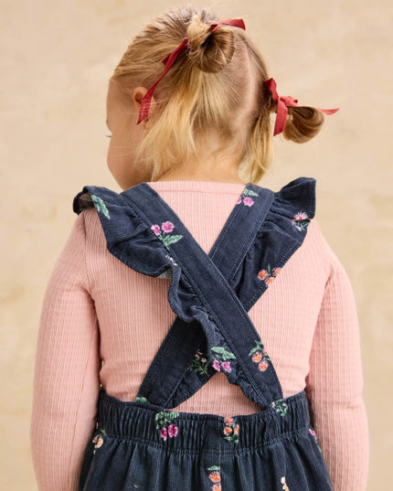 A toddler girl wearing deep midnight corduroy overalls with floral pattern and ruffle straps and a pink undershirt