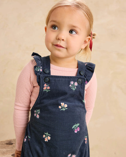 A toddler girl wearing deep midnight corduroy overalls with floral pattern and ruffle straps and a pink undershirt