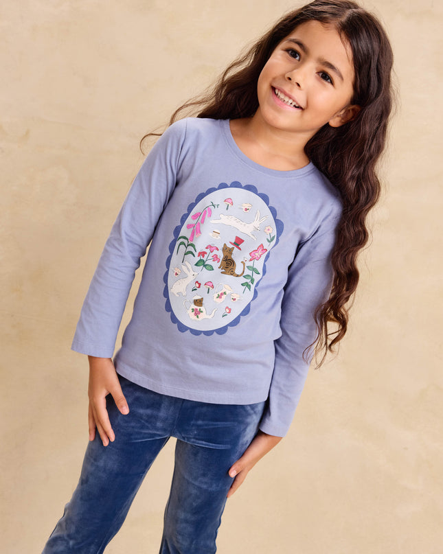 A young girl wearing a light periwinkle blue long-sleeve t-shirt with a colorful  tea party design on the front, paired with blue jeans
