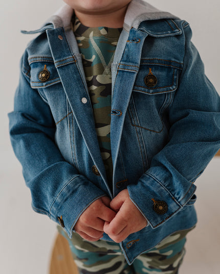 A toddler wearing a hooded blue denim jacket with a gray hood over a camouflage print top and matching camouflage pants