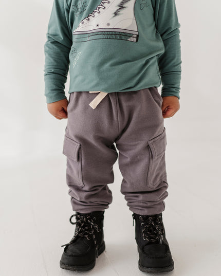 A child wearing a green long-sleeve shirt with a white sneaker graphic, gray cargo pants, and black boots