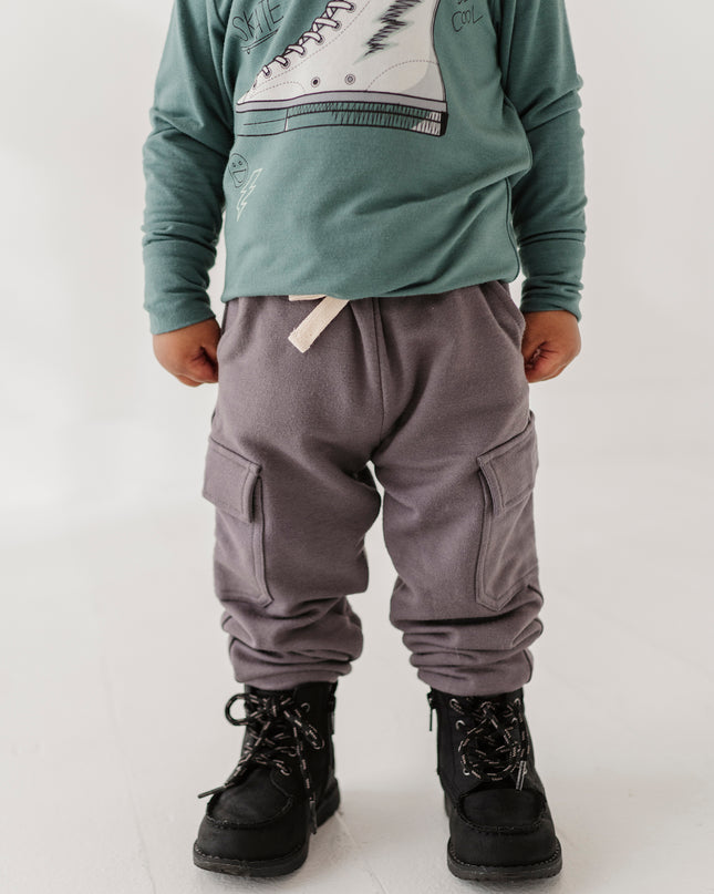 A child wearing a green long-sleeve shirt with a white sneaker graphic, gray cargo pants, and black boots