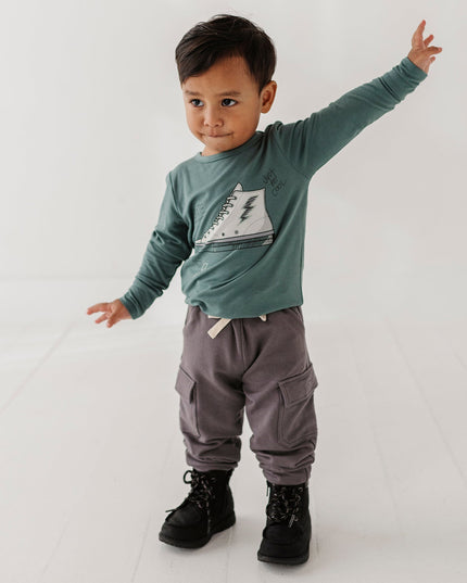 A child wearing a green long-sleeve shirt with a white sneaker graphic, gray cargo pants, and black boots