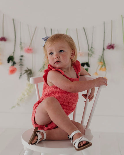 Baby in a red knit bodysuit sitting on a white chair with floral decorations in the background