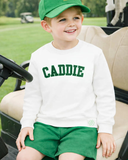 Child wearing a 'CADDIE' sweatshirt and green golf hat sitting in a golf cart 