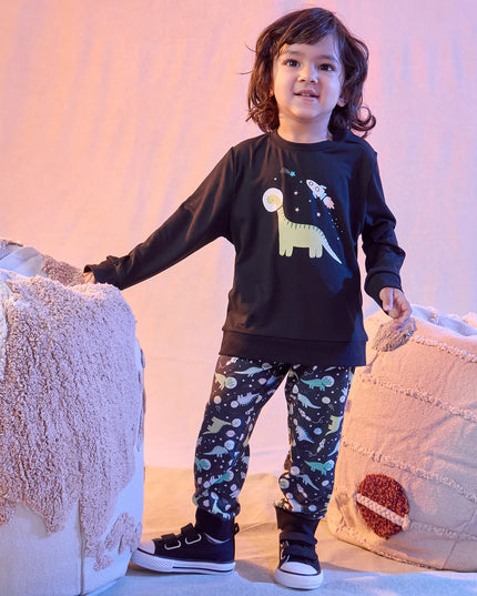 A boy wearing a two-piece kids lounge set featuring a black long-sleeve top with a dinosaur and space graphic, paired with black pants covered in a colorful dinosaur space print.