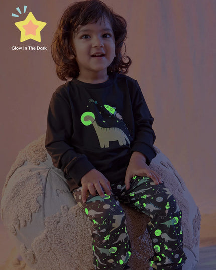 A boy wearing a two-piece kids lounge set featuring a black long-sleeve top with a dinosaur and space graphic, paired with black pants covered in a colorful dinosaur space print.
