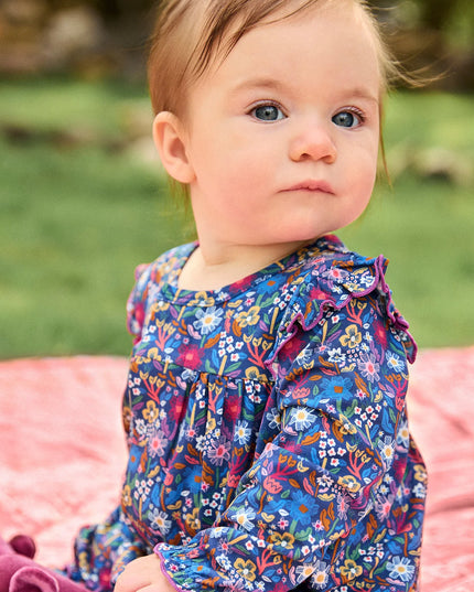 A baby girl wearing a long-sleeve floral romper, sitting on a pink blanket outdoors on a grassy lawn.