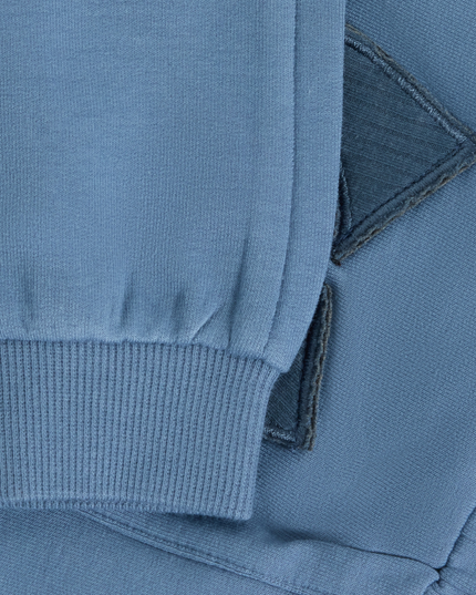 Close-up of a blue knit garment showing a ribbed cuff, folded fabric layers, and a dark contrast stitched detail.