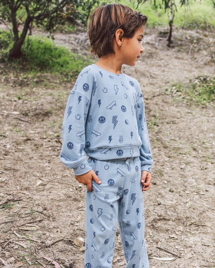 A young boy wearing a light blue long-sleeve sweatshirt and matching sweatpants  with a graphic print of lightning bolts, smiley faces, skateboards and peace signs