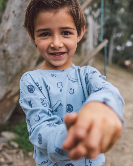 A young boy wearing a light blue long-sleeve sweatshirt and matching sweatpants  with a graphic print of lightning bolts, smiley faces, skateboards and peace signs