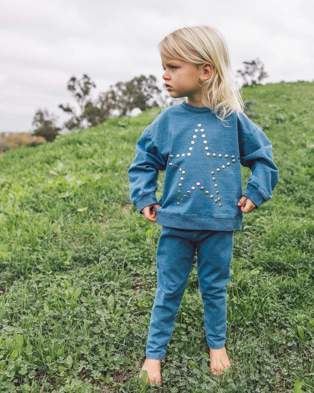 A young blonde girl wearing a matching blue sweatshirt and sweatpants set. The sweatshirt features a large star design outlined in silver studs.