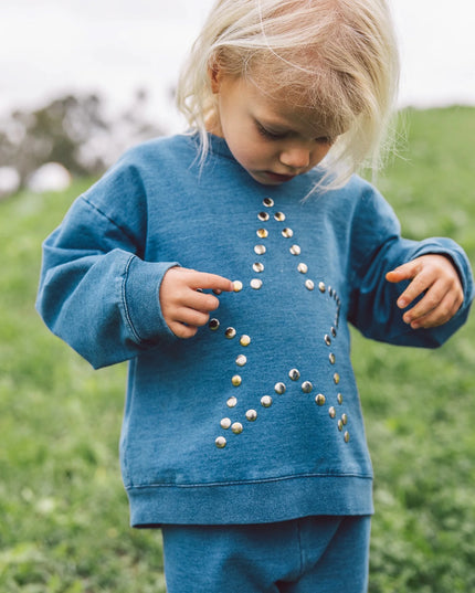A young blonde girl wearing a matching blue sweatshirt and sweatpants set. The sweatshirt features a large star design outlined in silver studs.