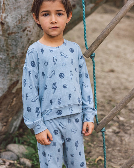 A young boy wearing a light blue long-sleeve sweatshirt and matching sweatpants  with a graphic print of lightning bolts, smiley faces, skateboards and peace signs