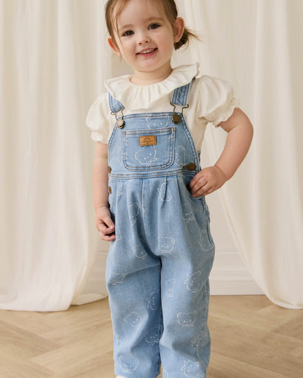 Child wearing blue denim overalls with bear pattern and white blouse