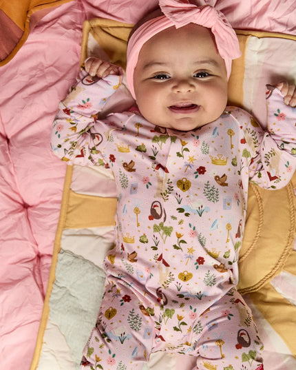 A baby wearing a convertible pink sleeper with a colorful woodland fairytale pattern of trees, flowers, and crowns, finished with pink trim on the collar.
