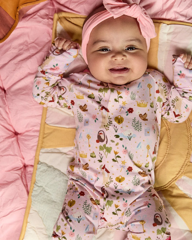 A baby wearing a convertible pink sleeper with a colorful woodland fairytale pattern of trees, flowers, and crowns, finished with pink trim on the collar.