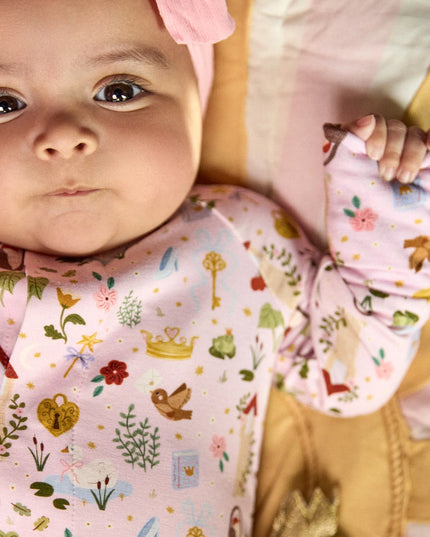 A baby wearing a convertible pink sleeper with a colorful woodland fairytale pattern of trees, flowers, and crowns, finished with pink trim on the collar.