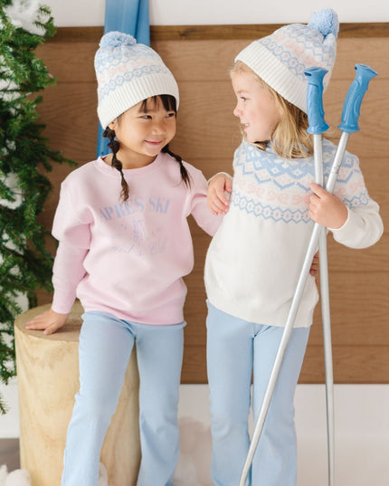A girl wearing a pink crewneck sweatshirt with blue apres ski club graphic and blue pants and another girl wearing a fair isle sweater and blue pants. 