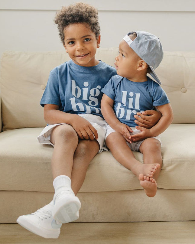 Two children sitting on a couch wearing matching big bro/lil bro blue shirts