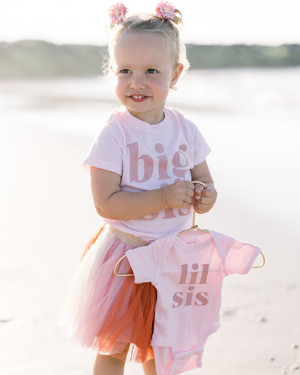 Child on a beach holding a 'lil sis' shirt, wearing a 'big sis' shirt.