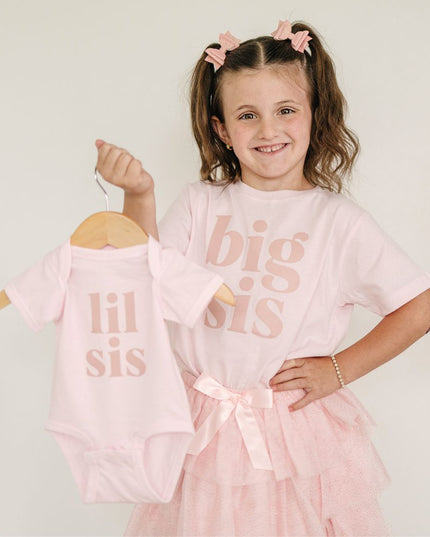 girl wearing a pink 'big sis' shirt with a matching tutu, holding a 'lil sis' outfit on a hanger.