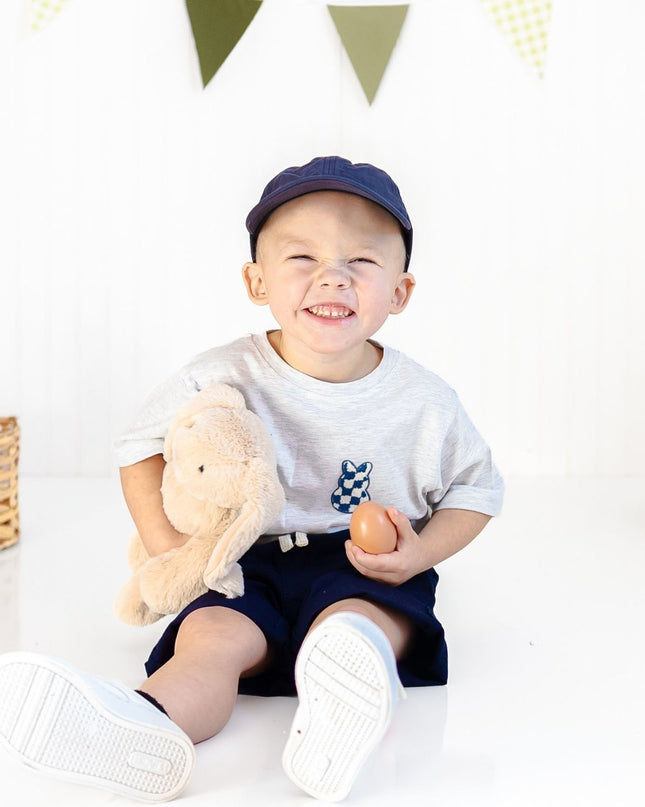 Boy wearing ash grey t-shirt with blue and white bunny checkered patch holding an egg and stuffed bunny