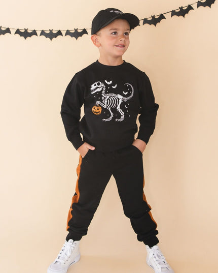A young boy modeling a black Halloween-themed two-piece sweatshirt and jogger set. The sweatshirt features a white skeleton dinosaur graphic with a small orange pumpkin, and the joggers have orange side stripes.