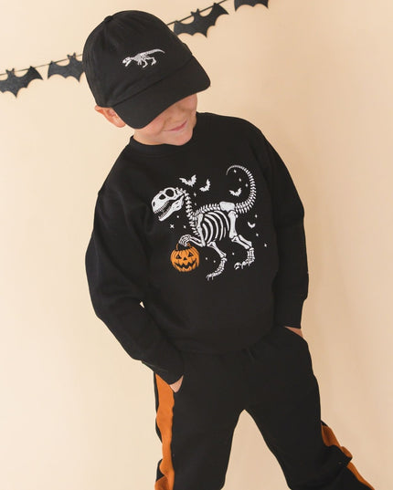 A young boy modeling a black Halloween-themed two-piece sweatshirt and jogger set. The sweatshirt features a white skeleton dinosaur graphic with a small orange pumpkin, and the joggers have orange side stripes.
