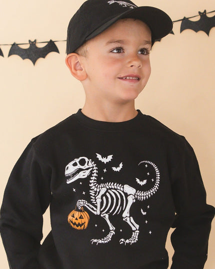 A young boy wearing a black long-sleeve crewneck sweatshirt with a white dinosaur skeleton and orange pumpkin graphic, paired with a black baseball cap