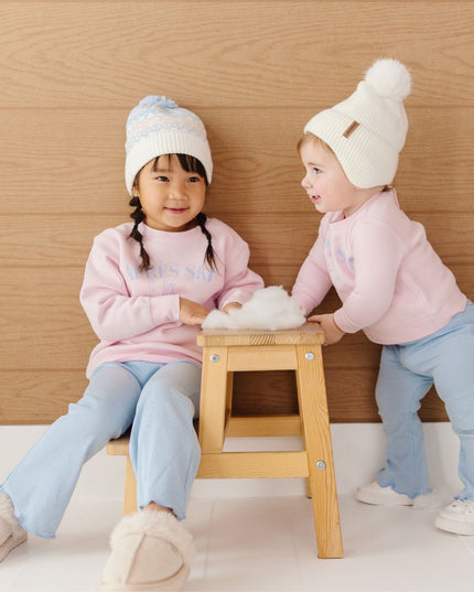 A girl and toddler wearing pink crewneck sweatshirts with blue apres ski club graphic, blue pants and beanies