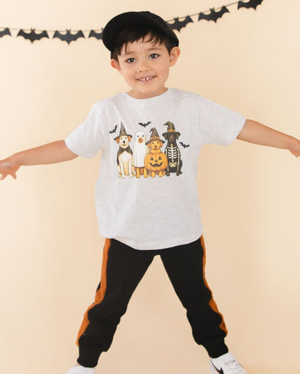 A young boy smiling and wearing a light gray heather t-shirt with a graphic of four dogs dressed in Halloween costumes and black joggers with orange side stripes