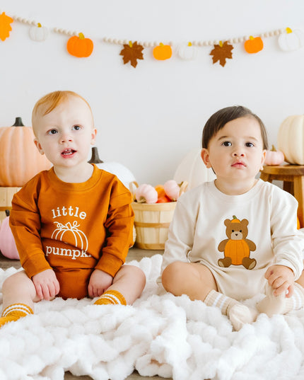 Two toddlers sitting on a white fluffy blanket in a fall-themed room wearing pumpkin outfits