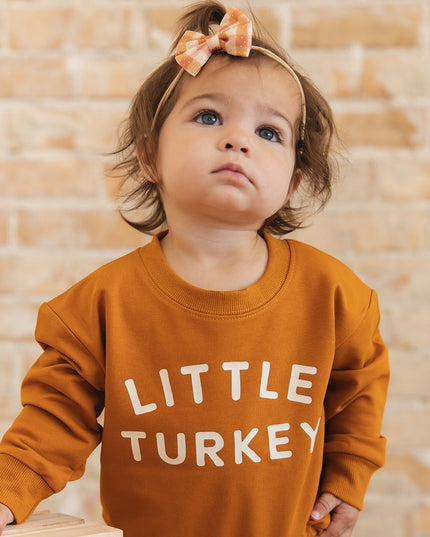 A baby wearing a burnt orange long sleeve romper with 'LITTLE TURKEY' printed in white