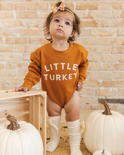 A baby standing wearing a burnt orange long sleeve romper with 'LITTLE TURKEY' printed in white
