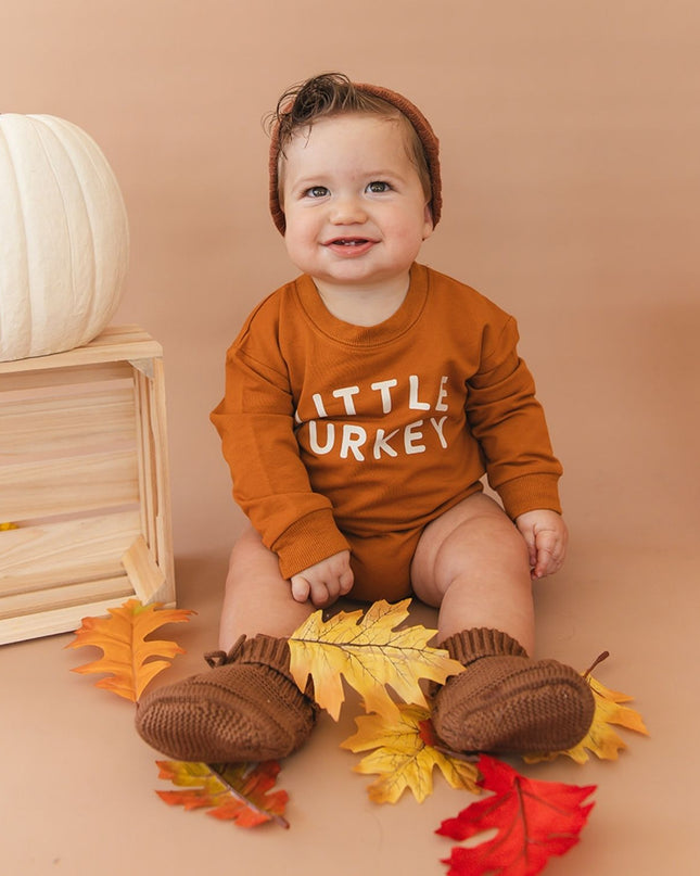 A baby sitting wearing a burnt orange long sleeve romper with 'LITTLE TURKEY' printed in white